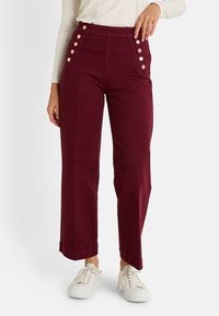 Pantalones burdeos de tiro alto con corte recto, que presentan seis botones visibles en el lateral y una textura de tela suave. Llevados con zapatillas blancas.