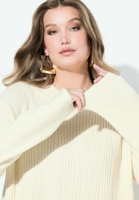 Krémový žebrovaný svetr s volným střihem a širokými rukávy. Modelka nosí velké zlaté náušnice ve tvaru kruhu, které zdůrazňují hladkou texturu.