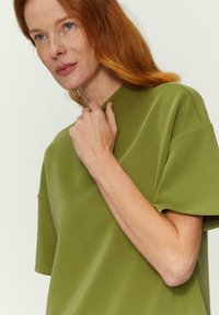 Olivenfarbiges, übergroßes T-Shirt mit hohem Halsausschnitt und kurzen Ärmeln. Sanfte Textur mit dezentem Glanz. Das Model hält den Kragen mit einer Hand.