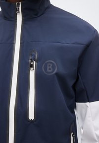Veste bleu marine avec des fermetures éclair blanches imperméables sur la poitrine et les côtés, dotée d'un col montant et d'un subtil logo circulaire « B ».