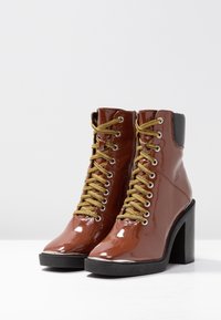 Bottines en cuir verni marron avec lacets dorés, talon noir et bout arrondi. Dotées d'une fermeture éclair latérale pour un enfilage facile.
