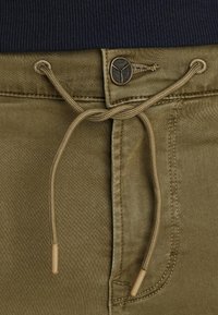Detail khaki kalhot ukazující šňůrku s dřevěnými konci, přiléhavý pás a kovový logo knoflík na texturovaném pozadí.