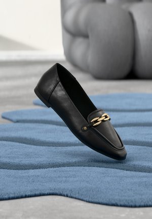 Mocassin en cuir noir avec détail chaîne dorée reposant sur un tapis bleu texturé, avec une chaise grise floue en arrière-plan.