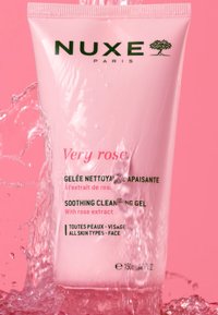 Rosa tub av NUXE Paris Soothing Cleansing Gel. Text i grönt och rosa. Vattensprut runt den, vilket framhäver dess släta yta och design.