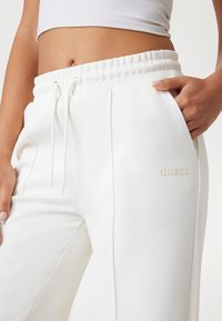 Witte sweatpants met een elastische tailleband en trekkoord, voorzien van zijzakken en een subtiel reliëf "GUESS" logo op de bovenbeen.
