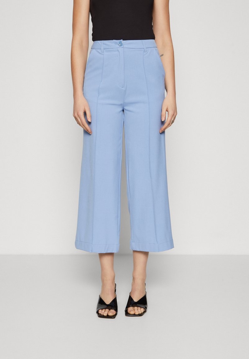 Femme portant un pantalon court large bleu clair avec des plis devant, un haut noir sans manches et des sandales à talons ouvertes noires.