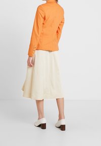 Oranssi istuva bleiseri, jossa on pitkät hihat ja kultaiset napit, yhdistettynä kermaiseen A-linjaisiin hameeseen. Asua täydentävät valkoiset korkeat kengät.