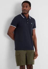Navy polo shirt made of cotton, featuring white collar and sleeve stripes, three-button placket, and a logo on the chest. Paired with khaki shorts.