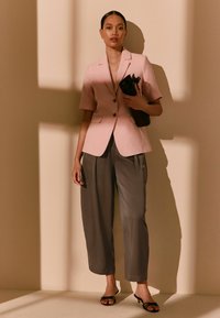 Femme debout en blazer rose clair à manches courtes, pantalon gris à jambes larges, talons noirs, tenant une pochette noire, devant un mur beige avec des ombres.