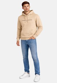 Giovane uomo con felpa beige con logo "Tommy Jeans", jeans azzurri e scarpe da ginnastica bianche, in piedi con una mano in tasca.