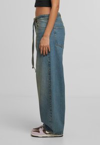 Osoba koja nosi dugu traper suknju visokog struka s bočnom džepom, crni kratki top i ružičaste i smeđe tenisice, stoji bočno.