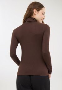 Donna con lungo capelli castani che indossa un dolcevita a maniche lunghe marrone, vista da dietro su uno sfondo bianco semplice.