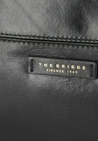 Borsa in pelle nera con una texture liscia. Presenta un'etichetta del marchio "THE BRIDGE" in lettere dorate e dettagli di cucitura visibili.