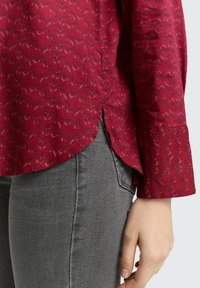 Rotes gemustertes Hemd aus strukturiertem Stoff mit wellenförmigem Design und seitlichen Schlitzen. Kombiniert mit grauen Jeans, die Passform und Stil betonen.