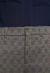 Detailní záběr na šedé a černé kostkované kalhoty s poutky na opasek, které jsou kombinovány s tmavě modrou košilí zasunutou dovnitř.