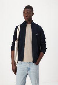 Marineblå zip-up genser i ribbestrikk, kombinert med en beige t-skjorte og lyseblå bukser. Modellen holder en kanvas tote bag. Enkelt design.