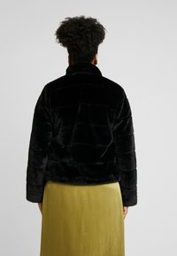Chaqueta de piel sintética negra con un cuello alto y un patrón texturizado acolchado. Llevada sobre una falda verde claro de longitud hasta la rodilla.