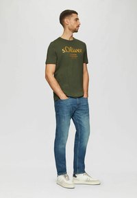 Mann steht mit den Händen in den Taschen, trägt ein olivgrünes s.Oliver T-Shirt, blaue Jeans und weiße Sneakers vor einem schlichten hellen Hintergrund.