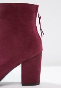 Bottine en daim bordeaux avec un talon haut épais et une fermeture éclair à l'arrière dotée d'une petite languette de tirage.