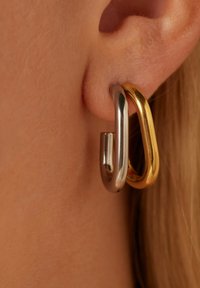 Twee met elkaar verbonden oorbellen: de ene zilver, de andere goud. Beide hebben gladde, afgeronde vormen en een glanzende afwerking, die dicht bij het oorlel hangen.