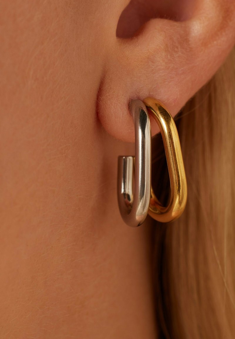 Twee met elkaar verbonden oorbellen: de ene zilver, de andere goud. Beide hebben gladde, afgeronde vormen en een glanzende afwerking, die dicht bij het oorlel hangen.