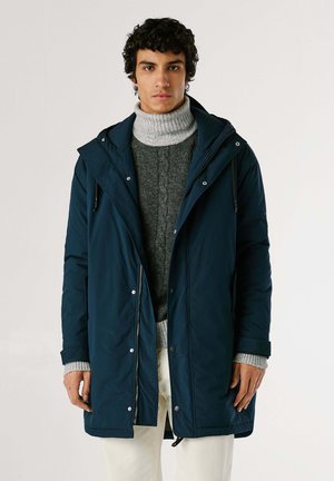 Abrigo acolchado azul marino con capucha, cierre de cremallera y botones a presión, y cintura con cordón. Llevado sobre un suéter de punto en gris y pantalones claros.