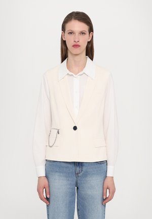 Lys rosa tettsittende vest med en enkelt knappelukking, to frontlommer og detaljer med kjede. Kombinert med en hvit skjorte og blå jeans.