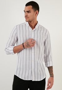 Camicia leggera in lino bianco con righe verticali nere, colletto button-down e maniche lunghe, con polsini arrotolati e vestibilità rilassata.