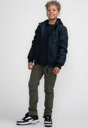 Giacca con cappuccio blu navy, pantaloni cargo verdi e sneakers bianche e nere. La giacca presenta una chiusura con zip e due tasche anteriori.