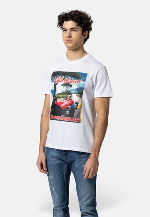 Jonge man met krullend haar die een wit t-shirt draagt met een rode raceauto en de tekst "Club Of Speed" en "The 19V69 Racing Bear."