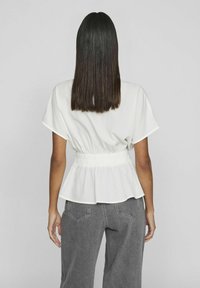 Top bianco a maniche corte con vita arricciata e design a peplo, abbinato a pantaloni grigi a gamba larga. Texture liscia con dettagli minimi.