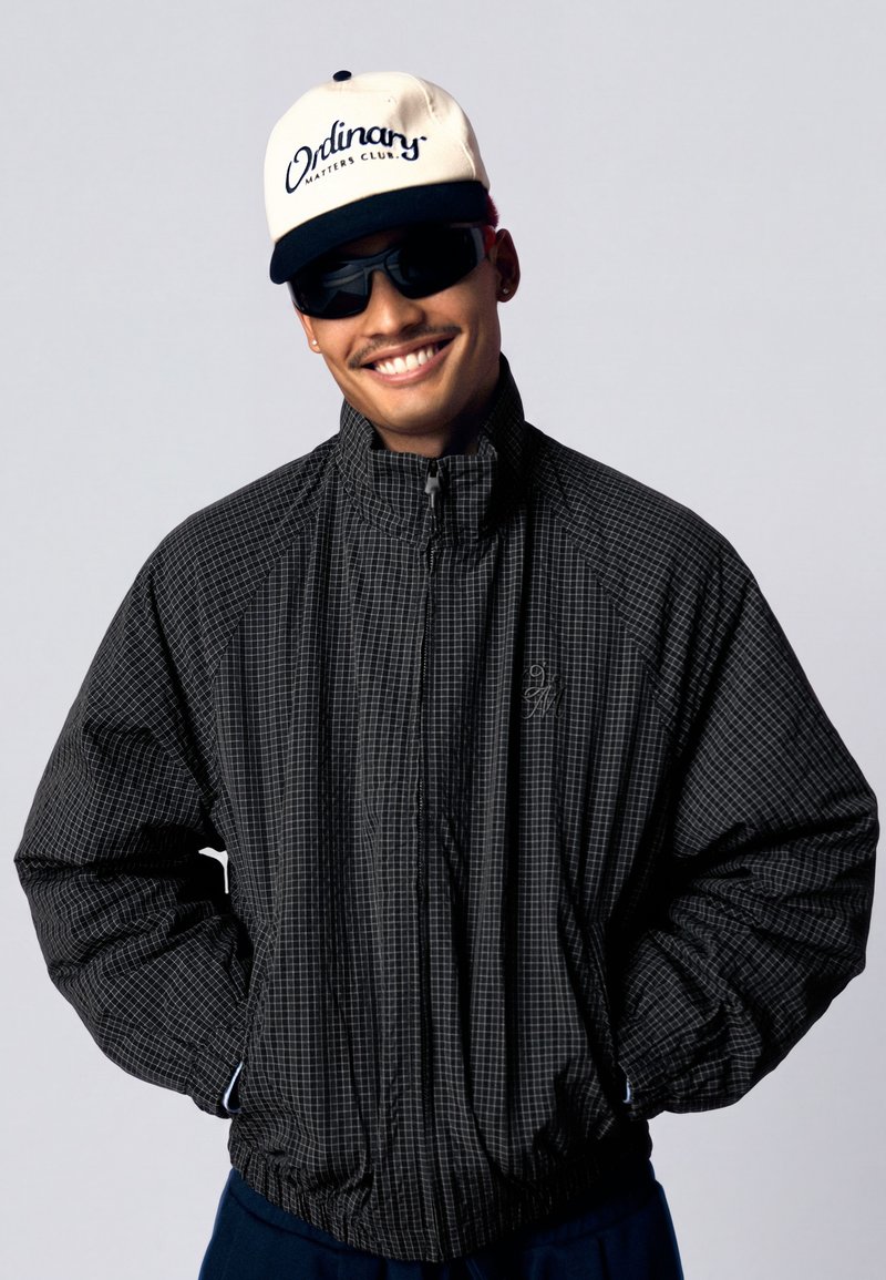 Homme souriant portant des lunettes de soleil, une casquette blanche avec "Ordinary Matters Club," et une veste noire à carreaux, les mains dans les poches, sur un fond clair.