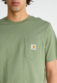 Oliivinvihreä lyhythihainen t-paita, joka on valmistettu puuvillasta ja jossa on rintatasku, jossa on Carharttin logo oranssina ja valkoisena. Miellyttävä, sileä pinta.