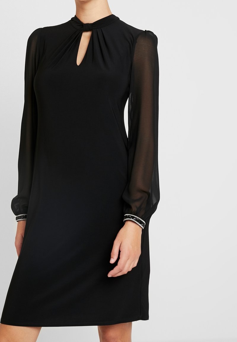 Schwarzes Kleid mit durchsichtigen langen Ärmeln, Ausschnitt mit Schlüsselöffnung und verzierten Manschetten, getragen von einer Person vor einem schlichten hellen Hintergrund.