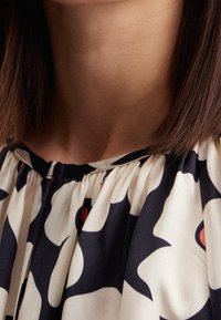 Blusa con colletto in crema con audaci motivi astratti neri e rossi. Presenta tessuto plissettato e una texture morbida e liscia.