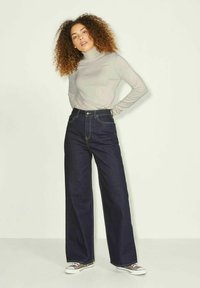 Jeans de mezclilla de pierna ancha en azul oscuro con cintura alta, complementados con un suéter de cuello alto de manga larga en gris claro y zapatillas grises.