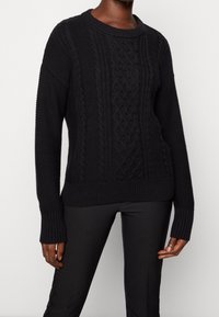 Mustassa neulotussa villapaidassa on teksturoitu palmikkokuvio, ribbaatut hihansuut ja helma. Pitkät hihat ja pyöreä pääntie viimeistelevät suunnittelun.