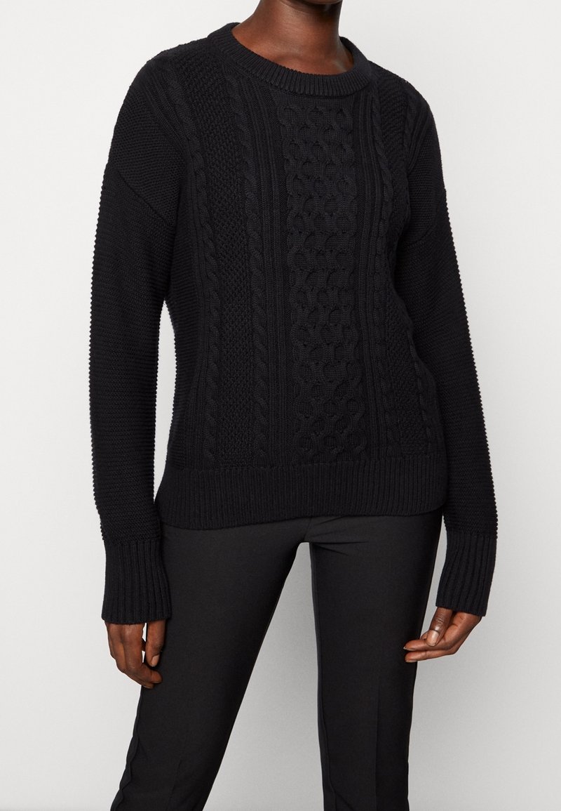 Mustassa neulotussa villapaidassa on teksturoitu palmikkokuvio, ribbaatut hihansuut ja helma. Pitkät hihat ja pyöreä pääntie viimeistelevät suunnittelun.