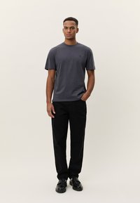 Vīriešu pelēks T-krekls no mīksta kokvilnas ar īsām piedurknēm, apaļu kakla izgriezumu un minimālistisku zīmolu. Kombinēts ar melnām biksēm un apaviem.