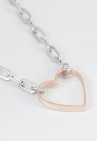 Zilveren schakelketting met een rosegouden hartpendant waarop het woord "LIEBE" is gegraveerd. Gladde metaaltextuur met een open ontwerp.