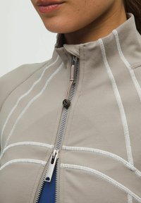 Jachetă zip-up de culoare gri deschis, cu detalii de cusătură albă. Material neted, guler înalt și elemente de fermoar argintii vizibile. Strat interior de culoare albastră.