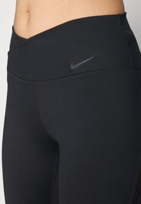 Leggings stretch noirs avec une taille haute, présentant une texture lisse et un petit logo Nike gris sur la zone de la taille avant.