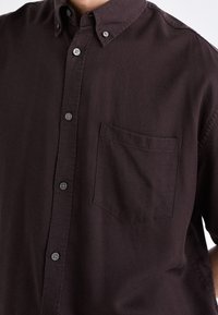 Donkerbruin kortärmelig overhemd met knopen, voorzien van een borstzak, een gladde textuur en een kraag met zwarte knopen als sluiting.