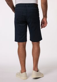 Námorníkově modré kraťasy vyrobené z bavlněné směsi, s dvěma zadními kapsami, prošitými švy a čistým střihem do délky kolen. Nosič s bílými teniskami.