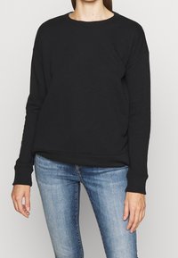 Černá mikina s kulatým výstřihem, spadlými rameny a žebrovanými manžetami. Spárovaná se světle modrými, přiléhavými džíny.