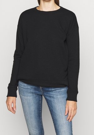 Felpa nera con scollo rotondo, spalle scese e polsini a costine. Abbinata a jeans attillati di colore blu chiaro.