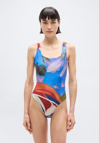 Γυναίκα με κοντά σκοτεινά μαλλιά, φορώντας μαγιό χωρίς μανίκια με πολύχρωμο αφηρημένο σχέδιο, στέκεται μπροστά σε απλό ανοιχτόχρωμο φόντο.