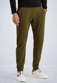Man in olijfgroene joggerbroek en witte sneakers, staand met één hand in zijn zak tegen een effen lichte achtergrond.