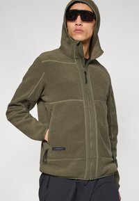 Μπουφάν με κουκούλα από φλις σε ελιά πράσινο χρώμα με τσέπες με φερμουάρ, αντιθετικές ραφές και απαλή υφή. Διαθέτει λογότυπο ραμμένο στο τελείωμα.