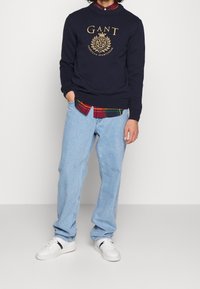 Granatowy sweter z dzianiny z złotym logo GANT, noszony na czerwono-zielonej koszuli w kratę, w połączeniu z jasnoniebieskimi dżinsami i białymi tenisówkami.